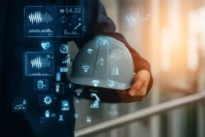Person holding a safety helmet with overlaid digital icons and data visualizations, representing industrial IoT with metrics for energy, connectivity, automation, health monitoring, and smart factory systems.